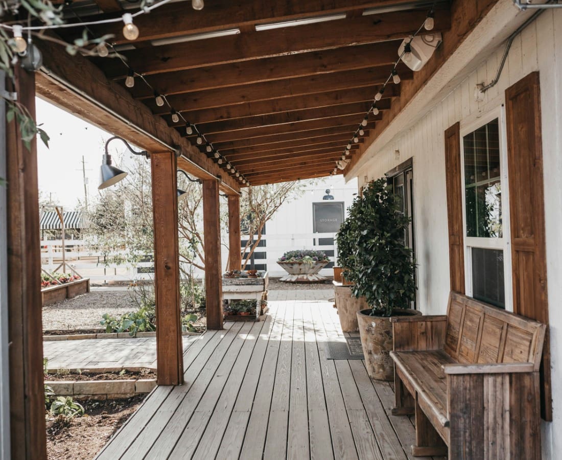 A wooden porch with string lights hangs from the ceiling. There is a bench on the right and potted plants along the edge. The ground is decorated with wooden planks and gravel. A building is visible in the background.