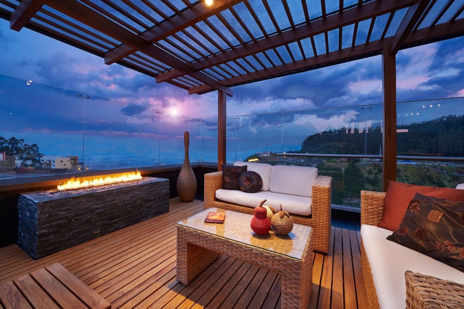 A rooftop patio with wicker furniture, a glass table, a long gas fire pit, and a wooden pergola. The area overlooks a scenic mountain view at dusk with a pink and purple sky.