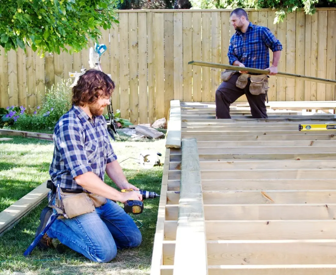 Two men in plaid shirts and tool belts work on constructing a wooden deck in a backyard. One is using a drill, while the other holds a plank.