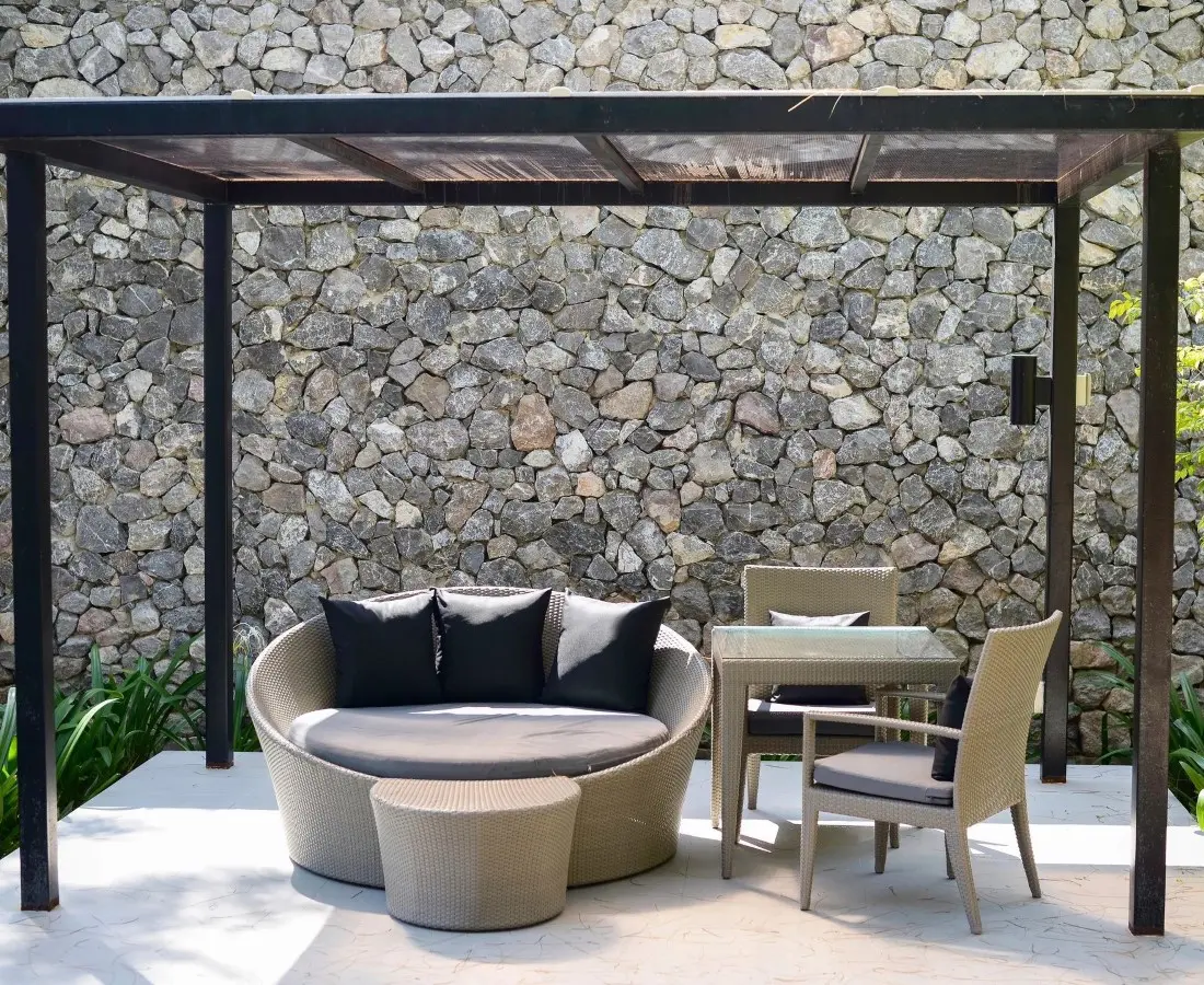 Outdoor seating area with a round woven sofa, black cushions, a matching ottoman, and a square glass-top table with three chairs under a pergola in front of a stone wall.