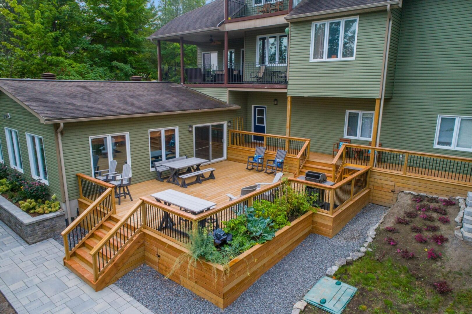 A multi-level deck attached to a green house features outdoor seating, planters, and a table. The house has multiple stories and is set in a landscaped yard with a stone walkway.