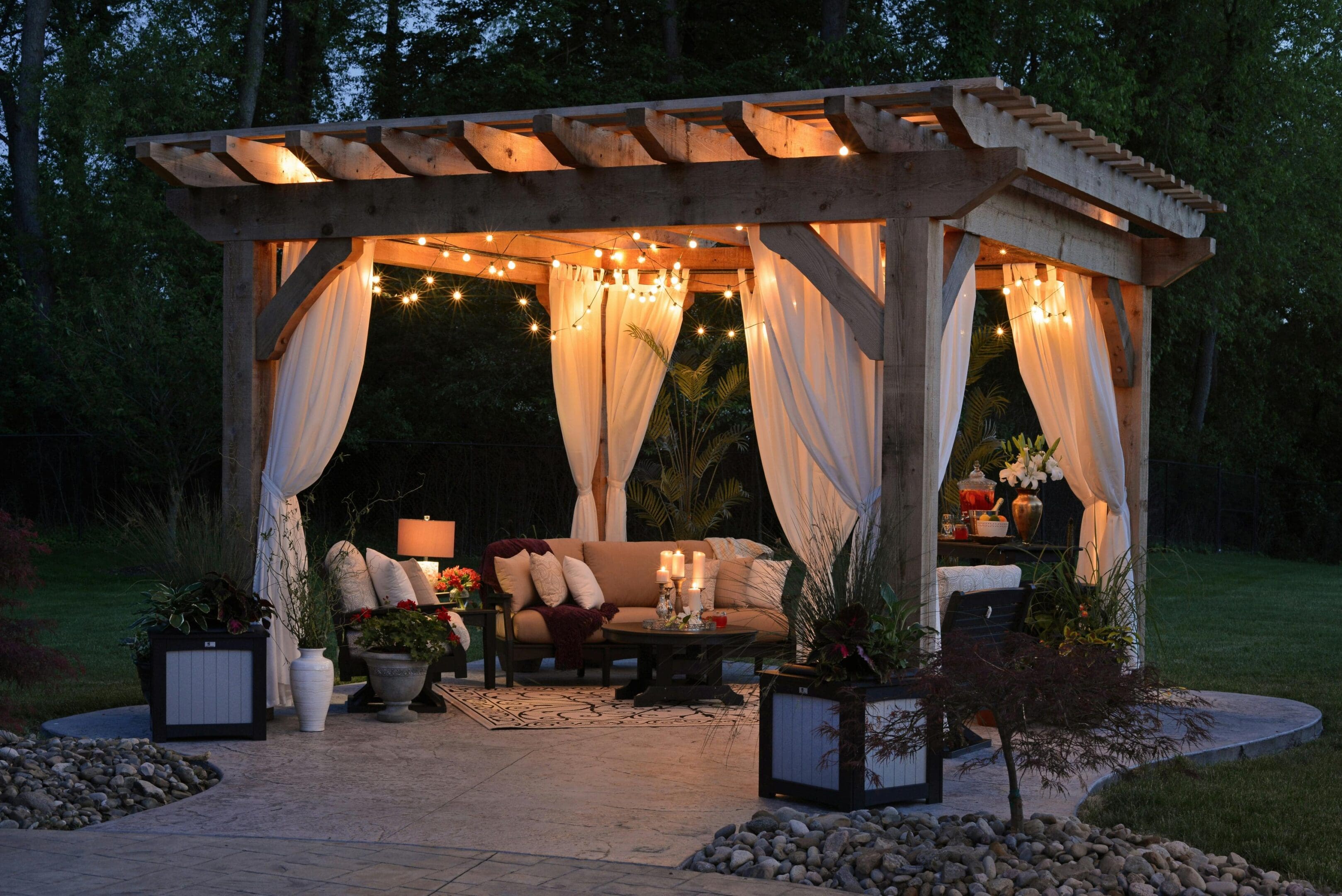 A wooden gazebo at night with glowing string lights, white curtains, and cozy seating inside, surrounded by plants and a pebble pathway.