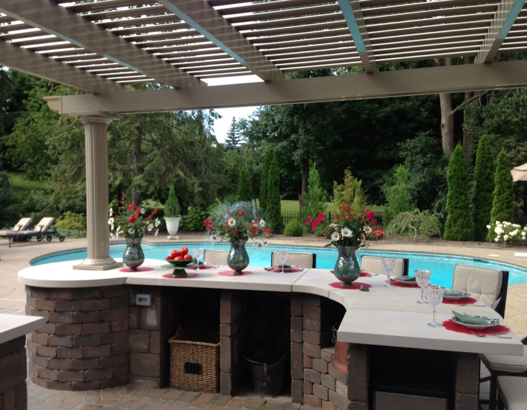 Elegant outdoor dining area under a louvered pergola, featuring a beautifully set table with floral arrangements, glassware, and a poolside view, enhancing outdoor living and relaxation.