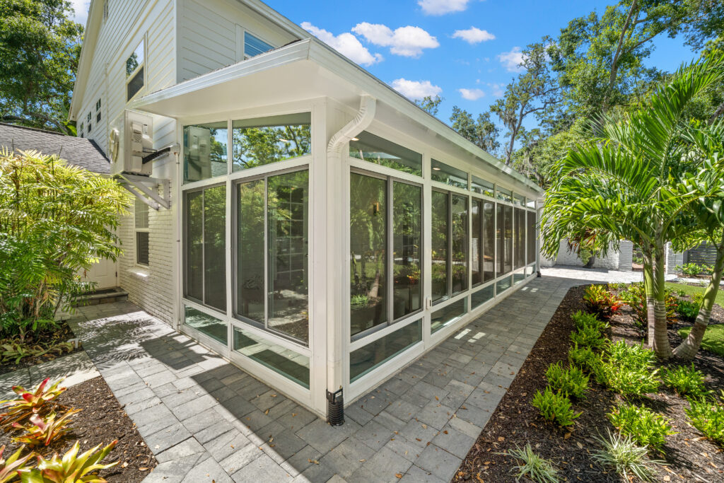 Custom sunroom with large glass windows, surrounded by lush greenery and colorful landscaping, showcasing an elegant outdoor living space designed for relaxation and enjoyment.