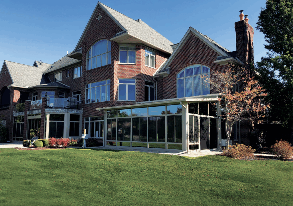 Elegant brick home with large windows and a custom sunroom featuring a glass enclosure, surrounded by a well-maintained lawn and landscaped garden, showcasing outdoor living solutions from Coastline Sunrooms & Awnings.